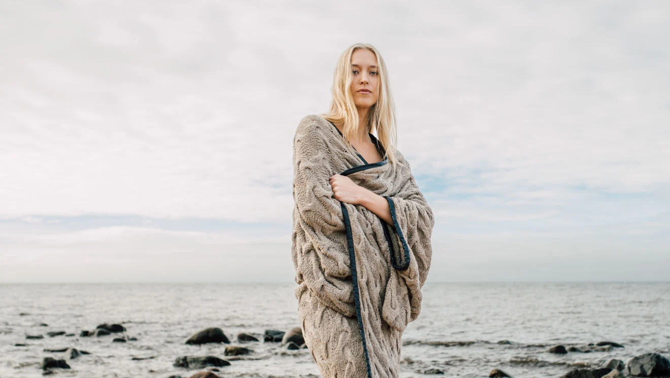 Woman wrapped in a knitted blanket on a Nordic pebble shoreline